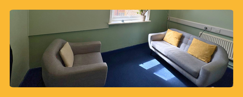 A modern counselling room in Islington with a grey sofa, yellow cushions, and a large window providing natural light.